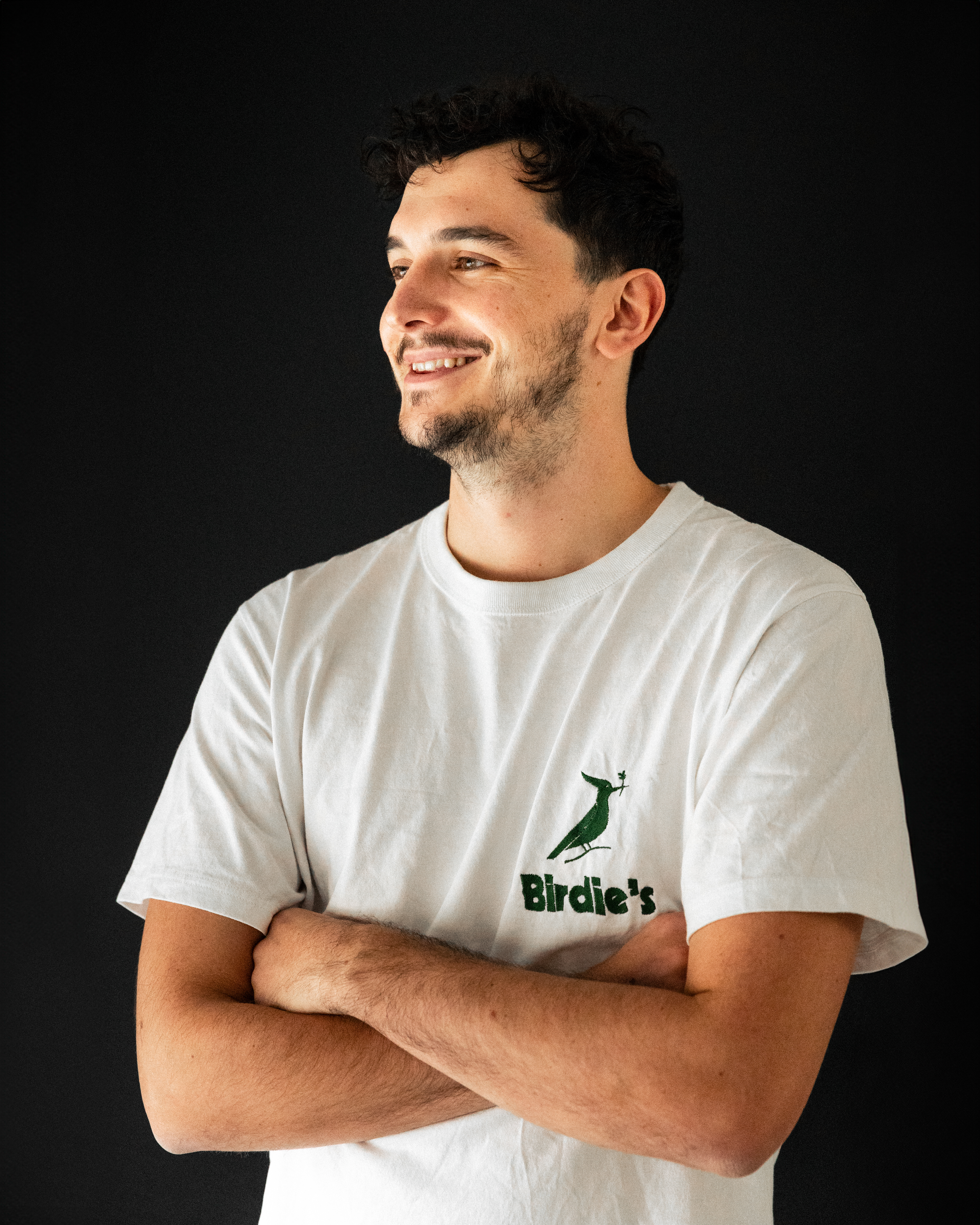 Une photo de Félix, un jeune homme brun habillé d'un tshirt blanc. Il pose de trois quart, les bras croisés, et souris à quelqu'un hors du champ de l'image.