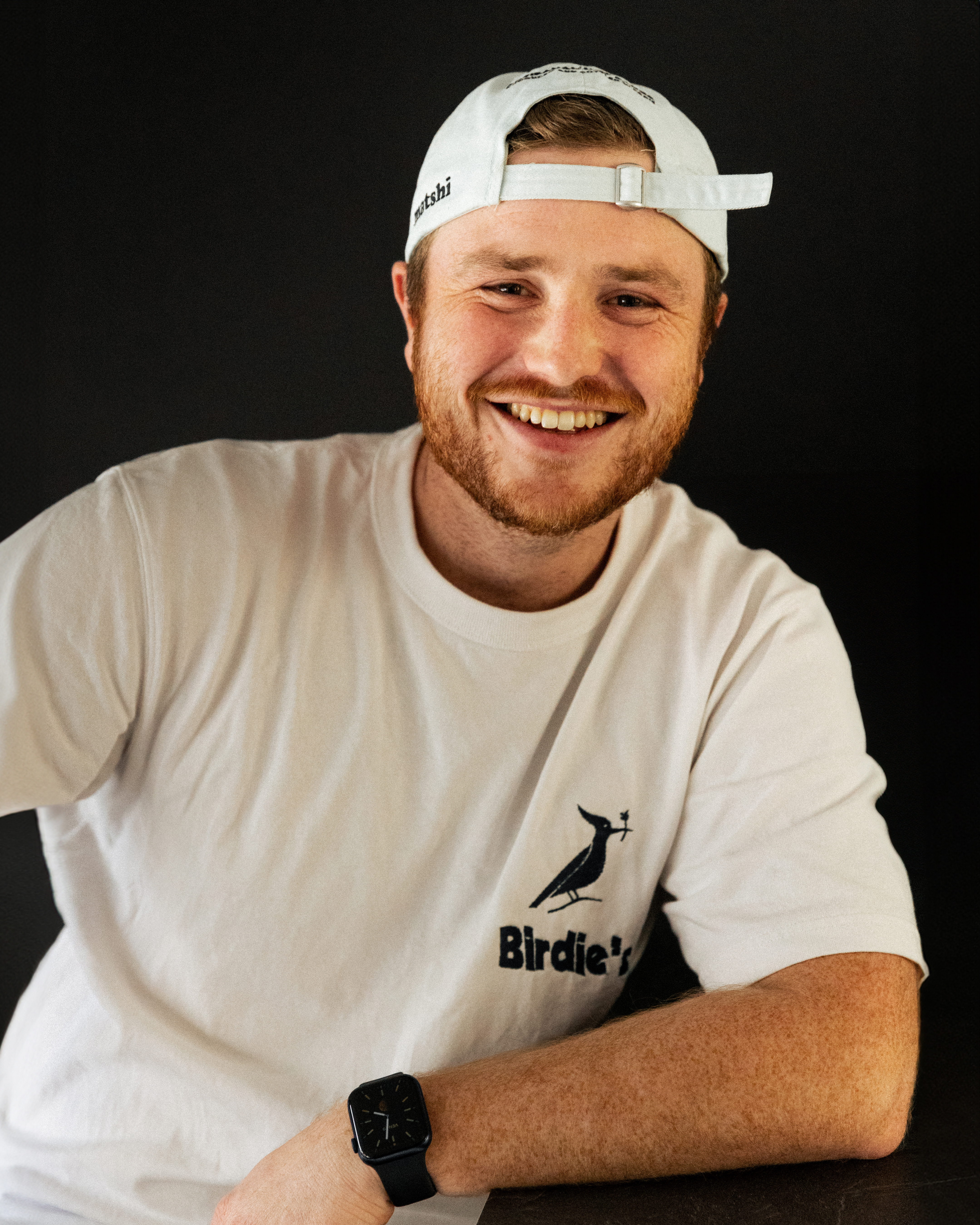 Une photo de Max, un jeune homme blond, vêtu d'un tshirt blanc et d'une casquette blanche. Il debout accoudé à une table et souris à la caméra.